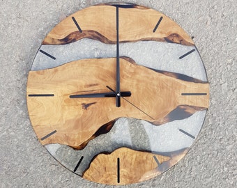 May include: A round wooden clock face with black hands and black Roman numeral markings. The wood has a natural grain pattern and is embedded with a gray and white epoxy resin that resembles a river.