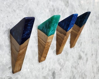 May include: Four wooden wall hooks with a geometric design. The hooks feature a natural wood base and a top section of resin in shades of blue and green. The hooks are mounted on a white textured wall.
