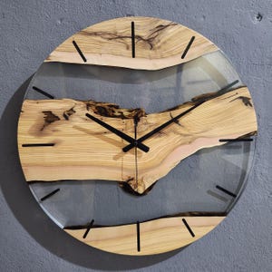 Puede incluir: Un reloj redondo de madera con una cara de resina epoxi transparente. La madera tiene un patrón de veta natural y las manecillas del reloj son negras.