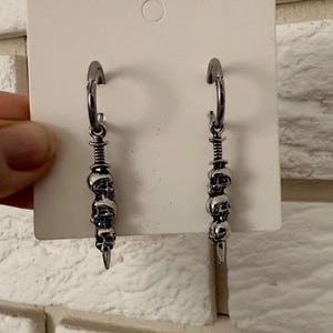 Puede incluir: Un par de pendientes de aro plateados. Cada pendiente presenta una serie de pequeños amuletos de calaveras suspendidos de un aro, que culminan en una punta afilada en forma de daga. Los pendientes se muestran sobre un fondo blanco.