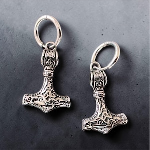 Könnte beinhalten: Zwei silberfarbene Anhänger in Form von Thors Hammer mit aufwendigen keltischen Knotenmustern. Jeder Hammer hat eine kreisförmige Schlaufe zum Aufhängen. Die Anhänger sind vor einem dunkelgrauen Hintergrund zu sehen.