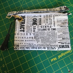 K&ouml;nnte beinhalten: Eine schwarze und wei&szlig;e Rei&szlig;verschlusstasche mit einem Zeitungsdruckdesign. Die Tasche hat einen schwarzen Quastenanh&auml;nger und einen goldenen Rei&szlig;verschluss-Zipper. Der Text auf der Tasche lautet "NEWS C. SMITH, the Cash Draper, Cuba Street EXP" und "TIMES SQ SHUFFLES - PARIS - ONE 15&cent;"