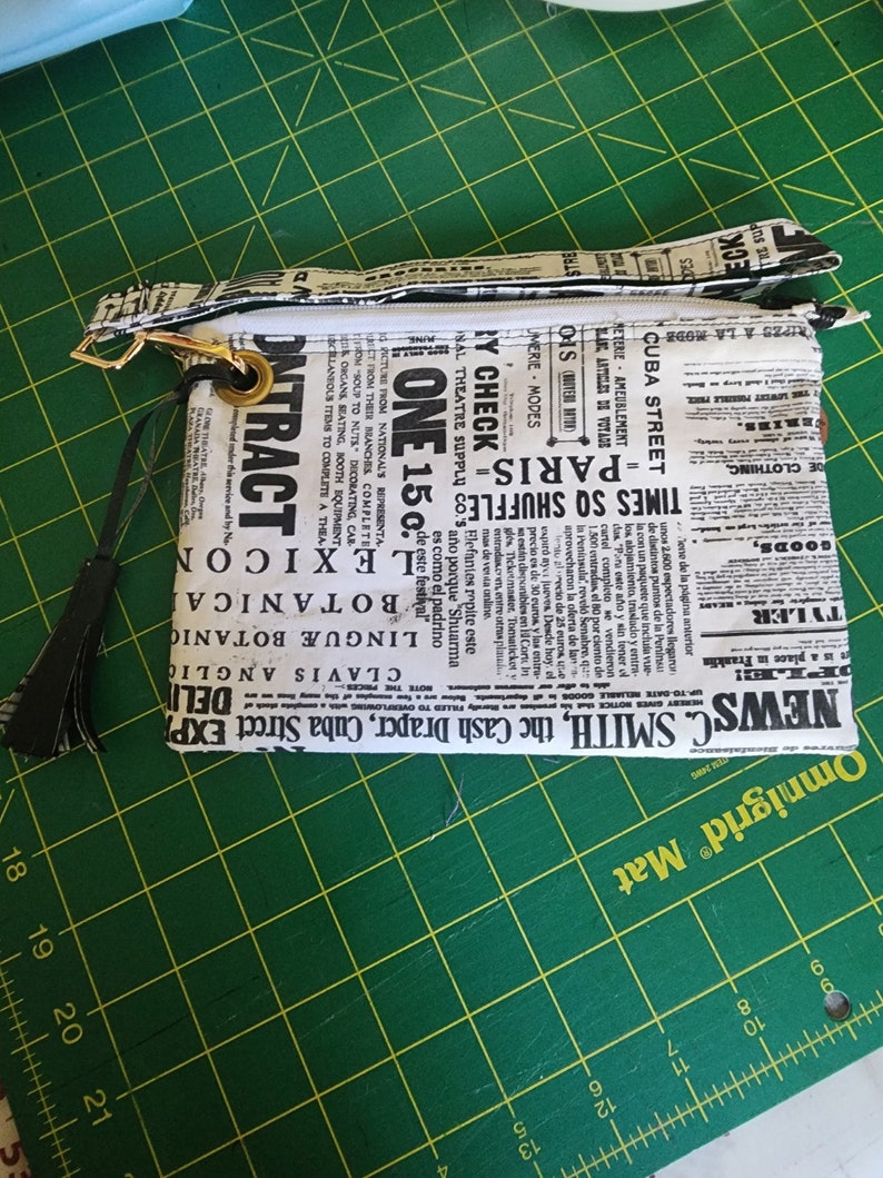 K&ouml;nnte beinhalten: Eine schwarze und wei&szlig;e Rei&szlig;verschlusstasche mit einem Zeitungsdruckdesign. Die Tasche hat einen schwarzen Quastenanh&auml;nger und einen goldenen Rei&szlig;verschluss-Zipper. Der Text auf der Tasche lautet "NEWS C. SMITH, the Cash Draper, Cuba Street EXP" und "TIMES SQ SHUFFLES - PARIS - ONE 15&cent;"