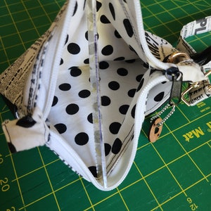 K&ouml;nnte beinhalten: Eine wei&szlig;e Rei&szlig;verschlusstasche mit schwarzem Punktmusterfutter. Die Tasche ist aus einem Stoff mit Zeitungsdruck. Die Tasche hat einen silbernen Rei&szlig;verschluss und eine silberne Kette, die an einem braunen Lederanh&auml;nger befestigt ist.