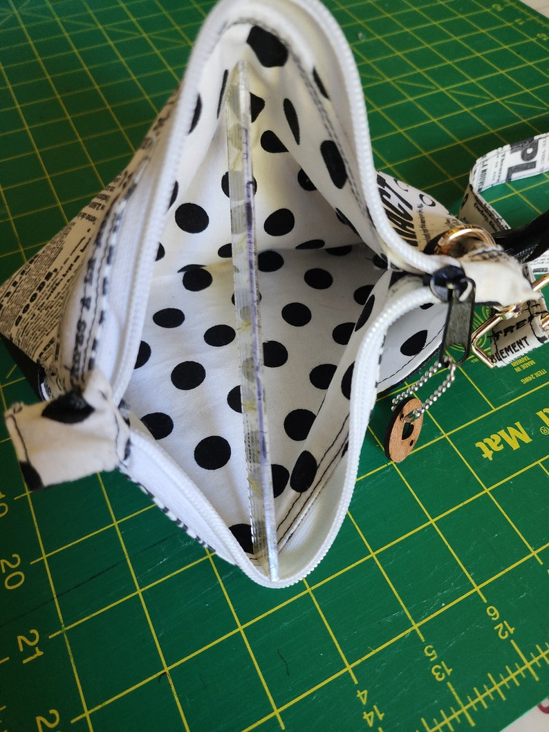 K&ouml;nnte beinhalten: Eine wei&szlig;e Rei&szlig;verschlusstasche mit schwarzem Punktmusterfutter. Die Tasche ist aus einem Stoff mit Zeitungsdruck. Die Tasche hat einen silbernen Rei&szlig;verschluss und eine silberne Kette, die an einem braunen Lederanh&auml;nger befestigt ist.