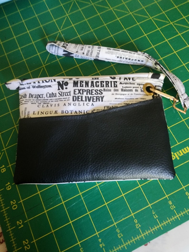 K&ouml;nnte beinhalten: Eine schwarze und wei&szlig;e Stofftasche mit einer schwarzen Lederklappe. Der Stoff hat einen Zeitungsdruck mit dem Text "No. Menagerie Express Delivery" und "Linguae Botanica". Die Tasche hat einen goldenen Rei&szlig;verschluss und einen schwarzen und wei&szlig;en Stoffgurt.
