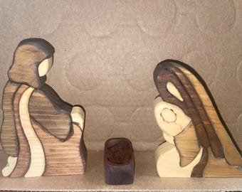 May include: A three-piece wooden nativity set featuring two stylized figures and a small central manger. The figures are crafted from contrasting light and dark wood grains, depicting two kneeling individuals facing a small, dark rectangular cradle.