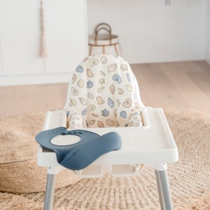 May include: A white high chair with a beige and blue seashell patterned seat cover. A blue silicone bib is on the tray.