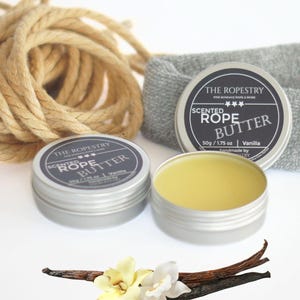 May include: An image of two silver tins of "Scented Rope Butter" with the text "The Ropestry" and "Vanilla". One tin is open, revealing the yellow butter. A bundle of rope, a gray cloth, and vanilla beans and flowers are also in the image.