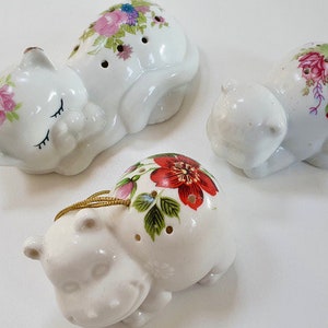 Puede incluir: Tres figuritas de cerámica, cada una representando un animal diferente. La primera es un gato blanco con diseños florales rosas y verdes, la segunda es un hipopótamo blanco con diseños florales rojos y verdes, y la tercera es un gato blanco con diseños florales rosas y verdes.