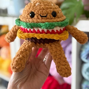 May include: A crocheted burger toy with a smiling face. The burger has a brown bun, green lettuce, red tomato, yellow cheese, and a brown patty. It has arms and legs made of textured yarn. The burger is held in a person's hand.