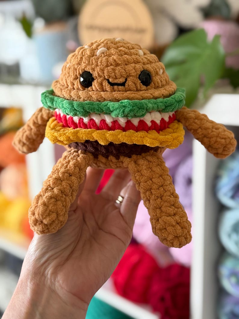 May include: A crocheted burger toy with a smiling face. The burger has a brown bun, green lettuce, red tomato, yellow cheese, and a brown patty. It has arms and legs made of textured yarn. The burger is held in a person's hand.