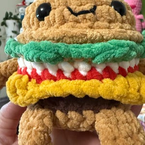 May include: A handmade, plush burger toy with a smiling face. The burger has a tan bun, green lettuce, white and red fillings, and a yellow bottom bun. Black eyes and a stitched mouth complete the design. The burger has arms and legs.