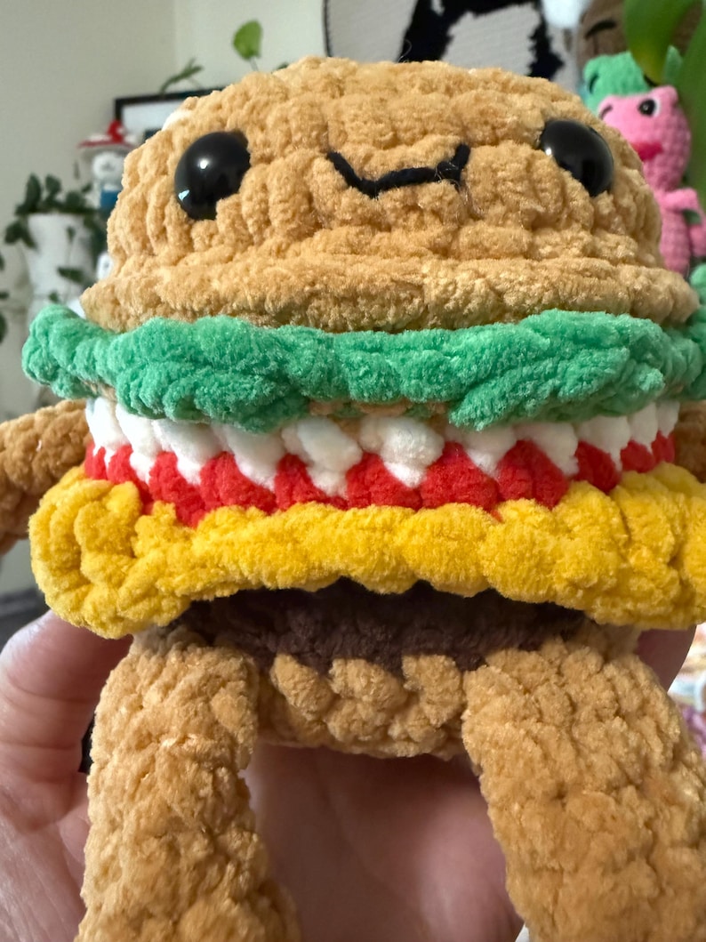 May include: A handmade, plush burger toy with a smiling face. The burger has a tan bun, green lettuce, white and red fillings, and a yellow bottom bun. Black eyes and a stitched mouth complete the design. The burger has arms and legs.