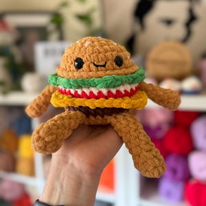 May include: A crocheted burger toy with a smiling face. The burger has a brown bun, green lettuce, red tomato, and yellow cheese. It has small black eyes and brown yarn arms and legs. A fun, whimsical food-themed toy, perfect for children or as a quirky gift.