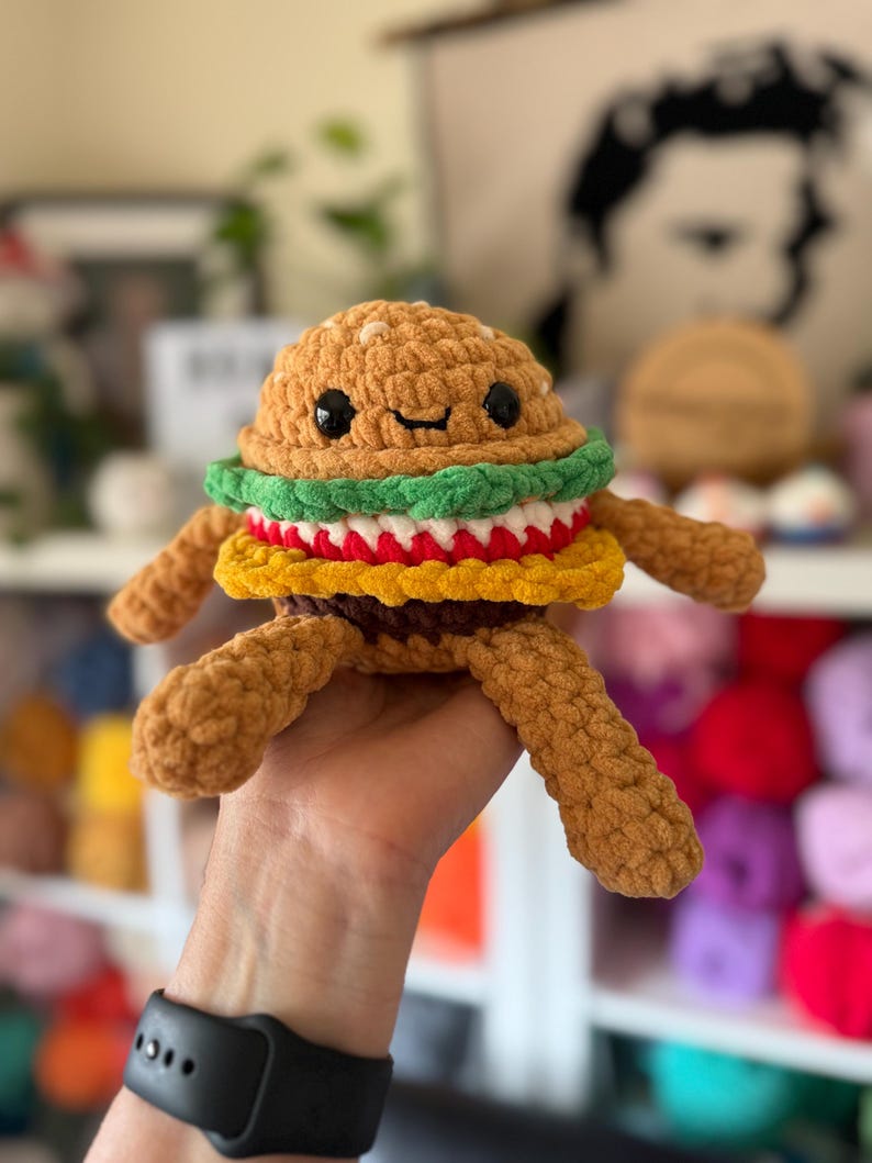 May include: A crocheted burger toy with a smiling face. The burger has a brown bun, green lettuce, red tomato, and yellow cheese. It has small black eyes and brown yarn arms and legs. A fun, whimsical food-themed toy, perfect for children or as a quirky gift.