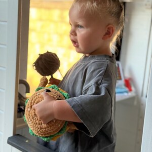 May include: A child holding a handmade crochet taco toy. The toy is made with brown, green, and yellow yarn, and has a smiling face. The child wears a grey t-shirt and has blonde hair tied in a bun.