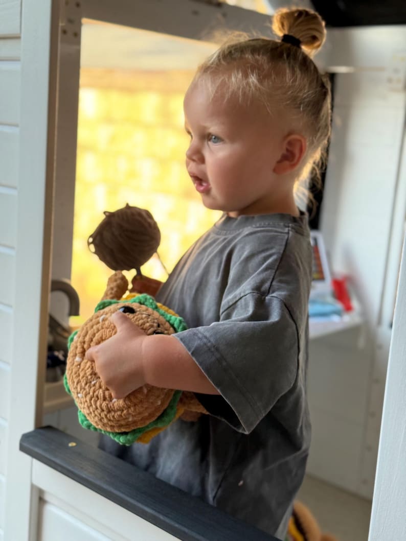 May include: A child holding a handmade crochet taco toy. The toy is made with brown, green, and yellow yarn, and has a smiling face. The child wears a grey t-shirt and has blonde hair tied in a bun.