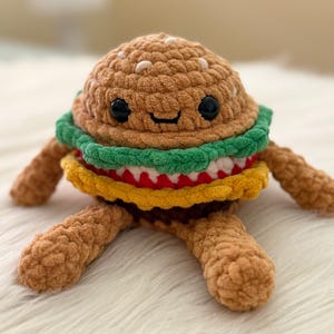 May include: A crocheted burger toy with a smiling face. The burger has a brown bun, green lettuce, red tomato, yellow cheese, and a brown patty. It has brown arms and legs, and is made of soft, textured yarn.