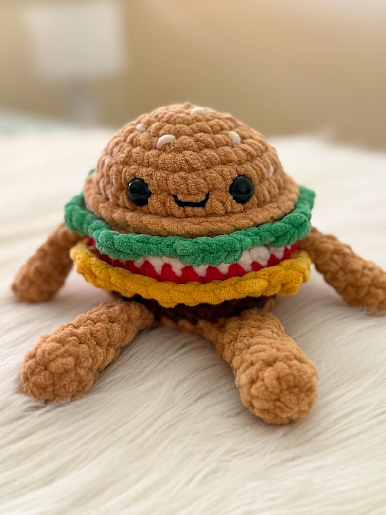 May include: A crocheted burger toy with a smiling face. The burger has a brown bun, green lettuce, red tomato, yellow cheese, and a brown patty. It has brown arms and legs, and is made of soft, textured yarn.