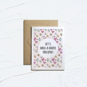 May include: A white card with a pink and blue seashell pattern. The card has a circle in the center with the text "Let's Shell-A-brate Our Love!"