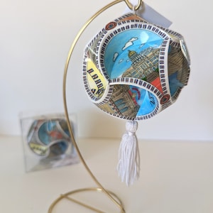 May include: A white globe ornament with a tassel, featuring a colourful illustration of a city skyline. The ornament is displayed on a gold metal stand.