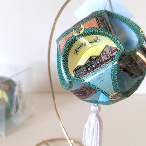 May include: A blue and green patterned ornament with a white tassel. The ornament is shaped like a cube and has a gold stand. The ornament features the text "Bowen's Wharf".