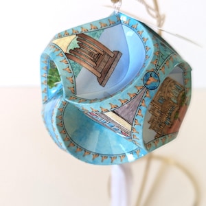 May include: A distinctive, geometric bauble with a light blue backdrop, featuring illustrations of buildings and landmarks. It hangs from a gold-coloured hook and white string. The design includes a repeating pattern of small, brown animal figures.