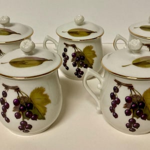 Puede incluir: Cinco tazas de porcelana blanca con tapas, cada una adornada con un borde dorado y un diseño pintado de uvas y hojas. Las tazas tienen asas y pequeños pomos en las tapas.
