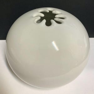 Może przedstawiać: Sferyczny, kremowobiały ceramiczny wazon z błyszczącym wykończeniem. Na górze znajduje się unikalny, stylizowany kwiatowy otwór. Wazon jest gładki i zaokrąglony, o prostym, eleganckim designie.
