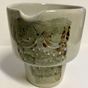 Puede incluir: Una crema de cerámica con un esmalte beige claro y un diseño de búho estilizado en tonos verdes y marrones. La crema tiene un pico y una base de pedestal, mostrando una estética rústica y hecha a mano. Los ojos y las plumas del búho están detallados con acentos marrones más oscuros.