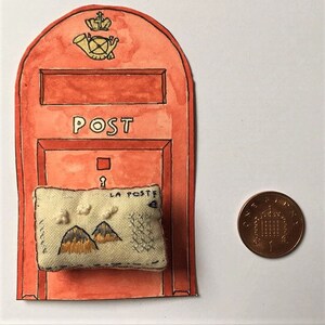 May include: A small, hand-stitched fabric postcard with a mountain scene and the words "La Poste" is placed in front of a red painted mailbox with the word "POST" on it. The mailbox has a small keyhole and a crown with a horn on top.