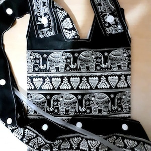 May include: Black and white patterned tote bag featuring an elephant motif. The bag has a shoulder strap and white button closures. The design includes rows of elephants and geometric shapes. The bag is made of fabric.