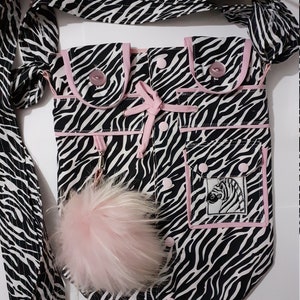 May include: A black and white zebra print crossbody bag with pink trim and buttons. The bag has multiple pockets, a pink fluffy pom-pom, and a strap. An embroidered zebra is on one pocket.