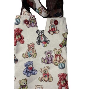 Puede incluir: Un bolso tote crema con un patrón repetido de osos de peluche coloridos. El bolso tiene un asa marrón y un borde superior decorativo. Los osos están en varias poses y colores, incluyendo rojo, azul y verde.