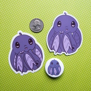 May include: Three purple bat-like creature stickers with big eyes and a happy expression. The stickers are on a green polka dot background. The largest sticker is about the size of a quarter.