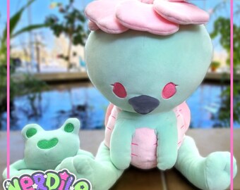 May include: A green plush toy with a pink flower on its head. The toy is sitting on a wooden surface. The toy has a small green frog plush toy in its lap. The frog plush toy has a happy expression.