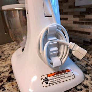 May include: White stand mixer with a silver bowl and a white cord wrapped around the base. The cord is secured with a white cord wrap. A warning label is visible on the base of the mixer that reads: "AWARNING Electrical Shock Hazard Unplug before servicing. Failure to do so can result in death or electrical shock."