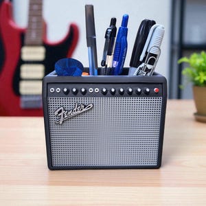 May include: A black desk organizer shaped like a guitar amplifier, with the "Fender" logo on the front. The organizer is filled with pens, markers, and other office supplies. The background includes a red guitar.