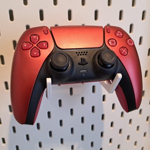 May include: A red PlayStation 5 DualSense wireless controller with black buttons and a white wall mount. The controller is mounted on a white pegboard.