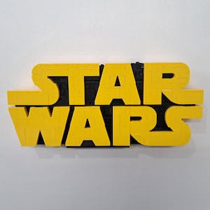 May include: A yellow and black "STAR WARS" sign made of interlocking blocks. The letters "STAR" are stacked above "WARS". The sign is mounted on a white wall.