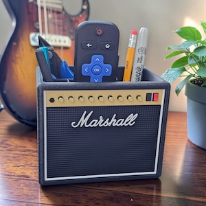 May include: A black desk organizer shaped like a Marshall guitar amplifier with a textured front grille, gold control knobs, and the white Marshall logo. The top compartment holds a black remote control, a yellow pencil, and a grey marker.
