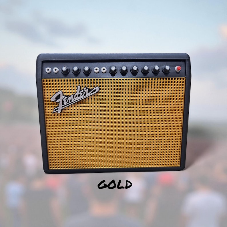 May include: A miniature gold and black Fender amplifier with the Fender logo. The amplifier has a gold mesh speaker cover and several knobs. The word "GOLD" is written in black script below the amplifier.