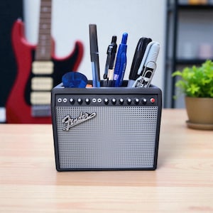 May include: A black desk organizer shaped like a Fender amplifier, with the Fender logo on the front. The organizer holds pens, markers, and a guitar pick. The background includes a red electric guitar and a potted plant.