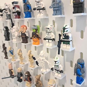 May include: A collection of Star Wars Lego minifigures displayed on a white pegboard. The figures are diverse in color and design, including characters in white, blue, green, and orange. Each figure is mounted on a small white shelf.