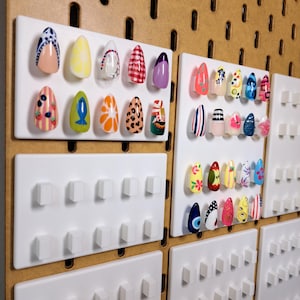 May include: A collection of colorful, decorated false nails displayed on white pegboards. The nails feature various designs, including fruit, animal prints, and geometric patterns. The background is a wooden pegboard.