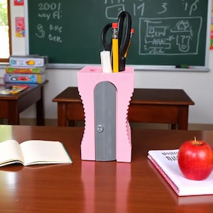 May include: A pink pencil sharpener-shaped desk organizer holds pencils and scissors. A red apple and an open book sit on the desk. A chalkboard with writing is in the background.