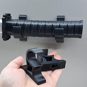 May include: Black plastic wall mount for a katana sword. The mount has multiple clips to hold the sword in place. A separate black plastic stand is also shown.