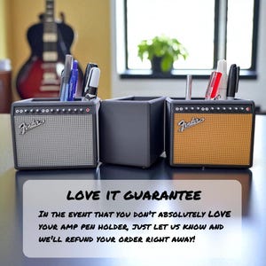 May include: Three pen holders designed to resemble guitar amplifiers. The pen holders are black with silver and orange speaker grilles. The Fender logo is visible on the front of the two amp-style holders. Pens and markers are inside the holders.