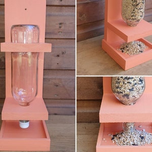 May include: A pink wooden bird feeder with two platforms. The top platform has a clear glass bottle for holding birdseed. The bottom platform has a tray for catching spilled seed.